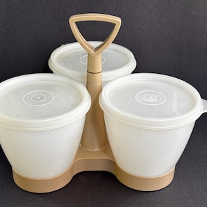 May include: A vintage Tupperware serving caddy with three white containers and lids. The caddy is beige with a handle for easy carrying. Each container has a lid with a circular design. The caddy is designed to hold condiments or snacks.