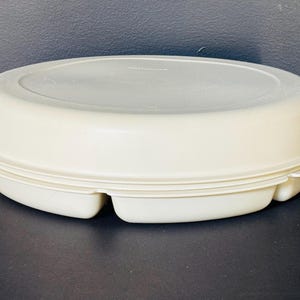 以下が含まれることがあります： オフホワイトの楕円形のタッパーウェア容器と蓋。容器には3つのコンパートメントに分かれたベースがあります。蓋はぴったりと収まり、密閉性を高めています。容器は耐久性のあるプラスチック製で、食品の保存用に設計されています。