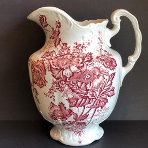 Könnte beinhalten: Eine weiße Keramikkanne mit rotem Blumendesign. Die Kanne hat eine breite Öffnung und einen Henkel.