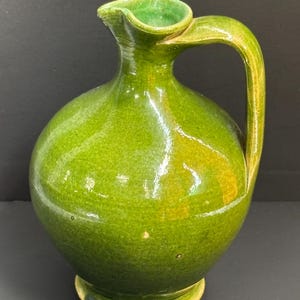 May include: A green ceramic pitcher with a rounded body, a spout, and a curved handle. The pitcher has a glossy finish and sits on a small, circular base. The interior of the spout is a lighter shade of green.