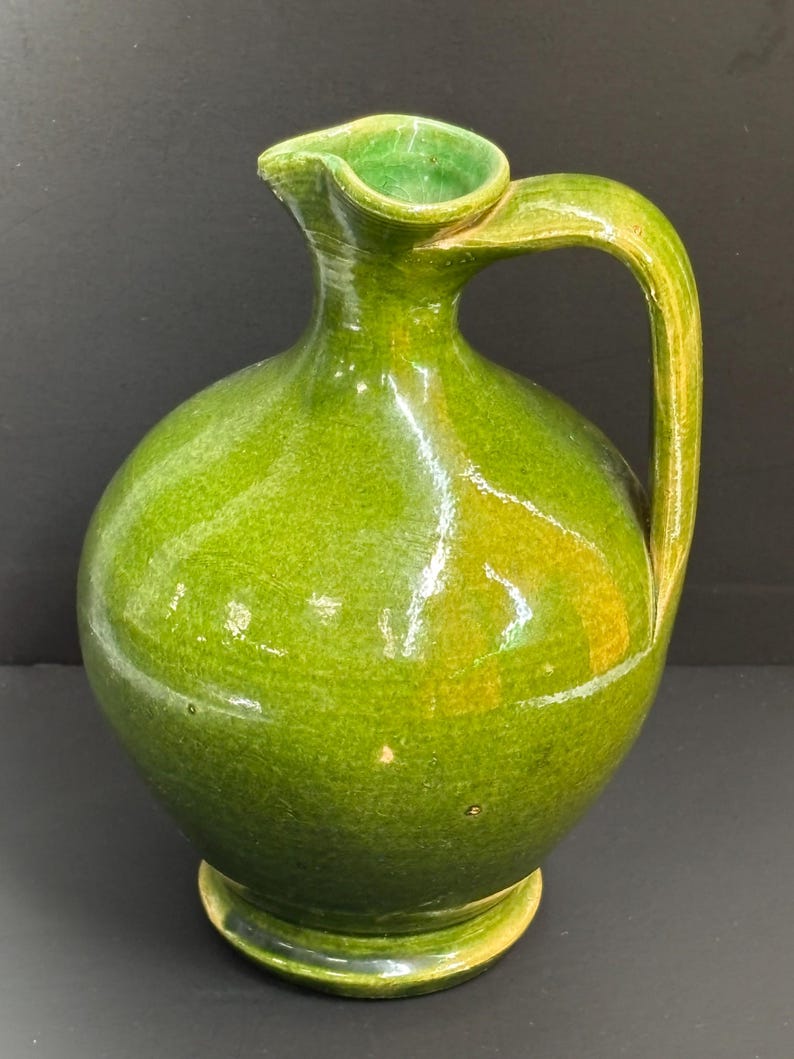 May include: A green ceramic pitcher with a rounded body, a spout, and a curved handle. The pitcher has a glossy finish and sits on a small, circular base. The interior of the spout is a lighter shade of green.