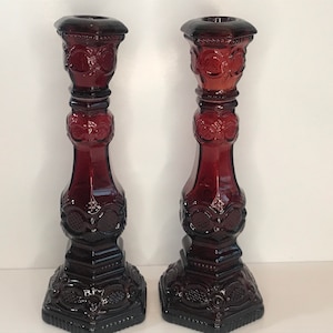 May include: A pair of dark red glass candle holders with intricate designs. The holders have a square base and a tall, slender body with a decorative top.