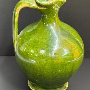 May include: A green ceramic pitcher with a rounded body, a curved handle, and a spout. The pitcher has a glossy finish and a base. The handle and spout have a lighter green color. The pitcher is likely used for pouring liquids.
