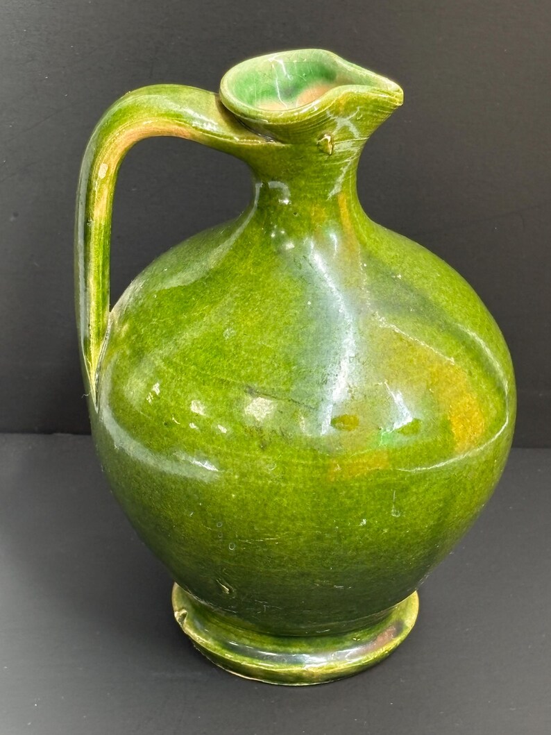 May include: A green ceramic pitcher with a rounded body, a curved handle, and a spout. The pitcher has a glossy finish and a base. The handle and spout have a lighter green color. The pitcher is likely used for pouring liquids.