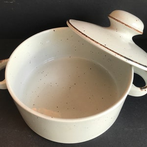Op de afbeelding: Een witte keramische ovenschaal met een gespikkelde glazuur en een bruine rand. De schaal heeft twee handvatten en een bijpassend deksel.