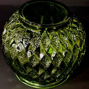 May include: A green glass vase with a diamond pattern. The vase has a wide, round body and a narrow opening. The glass has a textured surface that reflects light, creating highlights and shadows. The vase is likely used for decorative purposes.