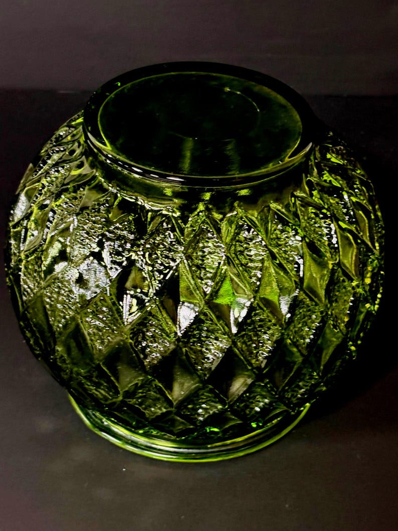 May include: A green glass vase with a diamond pattern. The vase has a wide, round body and a narrow opening. The glass has a textured surface that reflects light, creating highlights and shadows. The vase is likely used for decorative purposes.