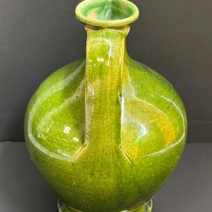 May include: A green ceramic jug with a rounded body, a handle, and a spout. The jug has a glossy finish and a base. The handle is attached to the body of the jug. The spout is a curved shape. The jug is a shade of green.