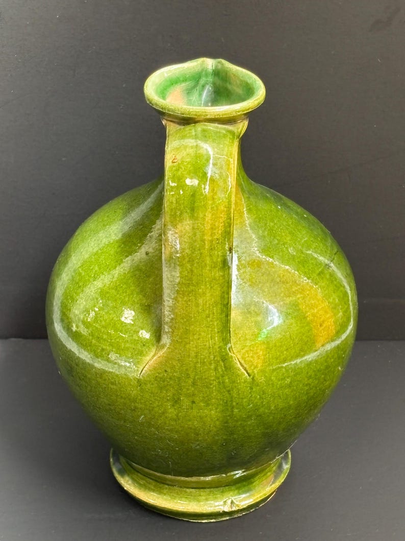 May include: A green ceramic jug with a rounded body, a handle, and a spout. The jug has a glossy finish and a base. The handle is attached to the body of the jug. The spout is a curved shape. The jug is a shade of green.