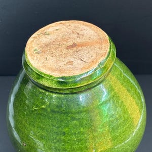 May include: A green glazed ceramic jar with a rounded body and a lid. The jar has a glossy finish with variations in the green color. The lid is a lighter brown color. The jar is likely an antique or vintage piece.