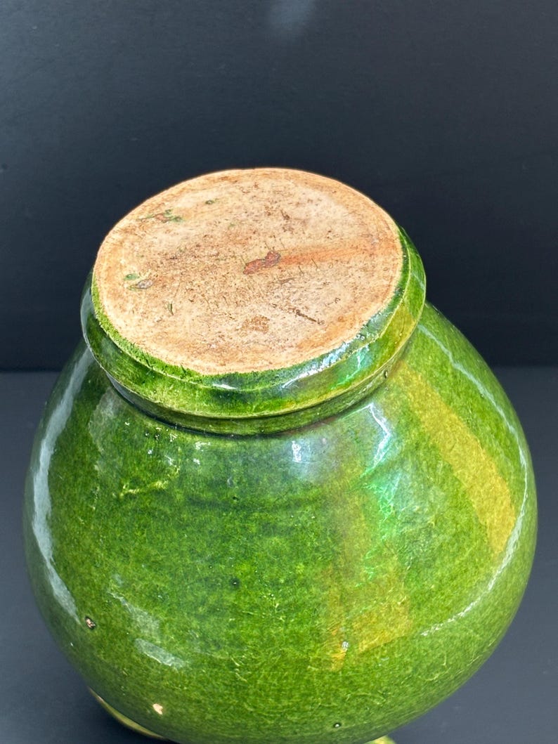 May include: A green glazed ceramic jar with a rounded body and a lid. The jar has a glossy finish with variations in the green color. The lid is a lighter brown color. The jar is likely an antique or vintage piece.