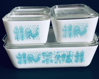 Puede incluir: Un juego de fuentes Pyrex blancas vintage con tapas de cristal transparente. Las fuentes presentan un diseño de gallo y flores turquesas. El juego incluye una fuente rectangular y tres recipientes cuadrados más pequeños, ideales para la cocina.