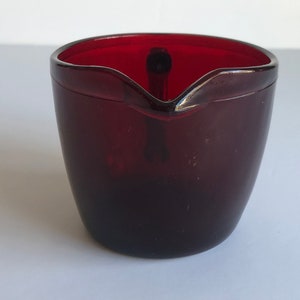 Puede incluir: Un pequeño creamer de vidrio rojo oscuro con asa. El creamer tiene una forma redondeada y un labio ligeramente acampanado.