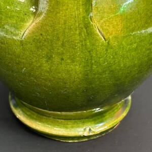 May include: Close-up of a green ceramic vase with a glossy finish. The vase has a rounded base and a wide, curved body. The green glaze has variations in color, with some areas appearing lighter and others darker. The vase appears to be handmade.