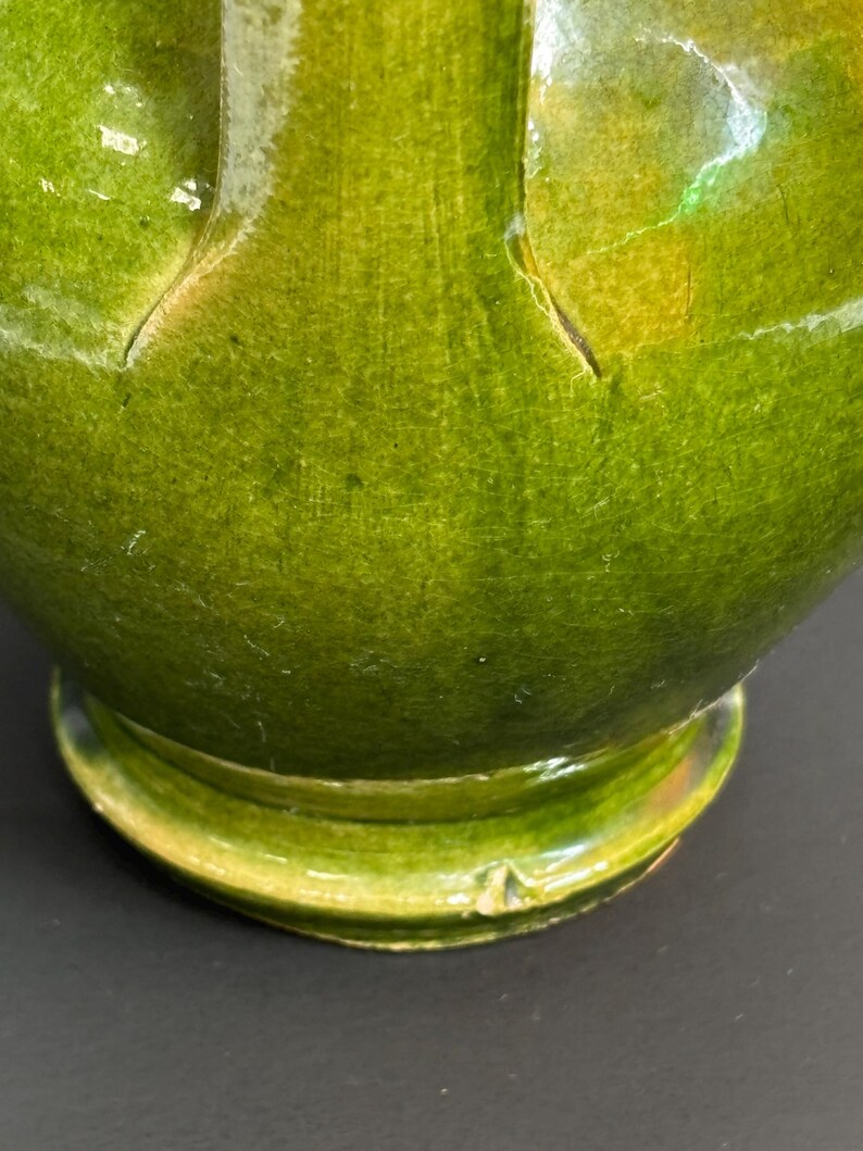 May include: Close-up of a green ceramic vase with a glossy finish. The vase has a rounded base and a wide, curved body. The green glaze has variations in color, with some areas appearing lighter and others darker. The vase appears to be handmade.