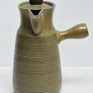 Peut inclure: Une cafetière en céramique vert olive. Le corps est texturé avec des rainures horizontales. Elle possède une anse incurvée, un bec verseur et un petit couvercle cylindrique. Design simple et fonctionnel, idéal pour le café.