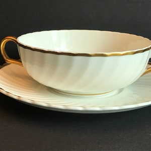Peut inclure: Bol de soupe en porcelaine blanche avec une bordure dorée et deux anses dorées. Le bol est posé sur une soucoupe assortie.