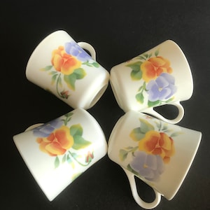 Puede incluir: Cuatro tazas de cerámica blanca con un diseño floral. Cada taza presenta un grupo de pensamientos amarillos, naranjas y morados con hojas verdes.