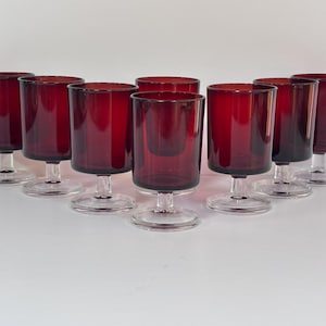 May include: A set of eight matching drinking glasses featuring deep ruby red bowls with straight sides. Each glass is supported by a clear glass stem and a circular clear glass base, creating a uniform appearance across the entire collection.