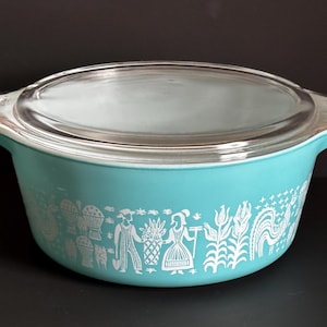 以下が含まれることがあります： 透明なガラス蓋付きのターコイズ色のパイレックスキャセロール皿。皿には、人、花、バスケットの白い様式化されたデザインが施されています。ハンドルはデザインに統合されており、蓋はぴったりと収まります。