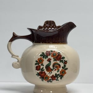 Könnte beinhalten: Cremefarbener Keramikkrug mit braunem Deckel und Griff. Ein kreisförmiges Design auf der Vorderseite zeigt einen Hahn, der von orangefarbenen und roten Blumen umgeben ist. Der Krug hat einen bauchigen Körper und einen Sockel.