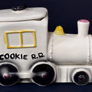 May include: A white ceramic cookie jar shaped like a steam locomotive train. The train features black wheels, a yellow window frame, a pink smokestack top, and the black text COOKIE R.R. printed on the side. It includes a removable lid.