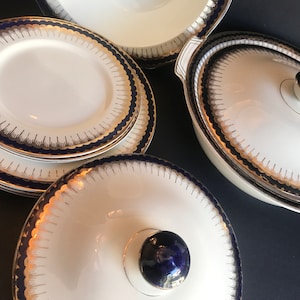 Puede incluir: Un juego de platos de porcelana blanca con ribetes azules y dorados. Los platos son redondos y tienen un borde festoneado. El juego incluye una fuente grande con tapa, una fuente más pequeña y dos platos.