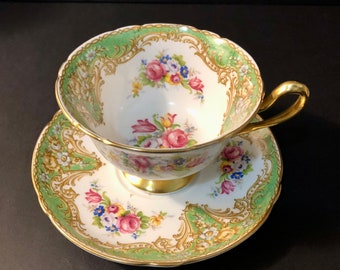 Peut inclure: Service à thé vintage composé d'une tasse et d'une soucoupe. La tasse et la soucoupe sont blanches avec une bordure verte et dorée. Elles présentent un motif floral avec des fleurs roses, bleues et jaunes. La tasse a une anse dorée et un pied doré.