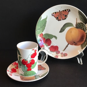 May include: A white porcelain teacup and saucer set with a floral design featuring red raspberries, green leaves, and a butterfly. The cup and saucer are on a black background.