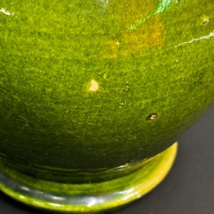 May include: A close-up of a green ceramic bowl with a glossy finish. The bowl has a rounded shape with a yellow rim and a dark base. The green color has variations, with some areas appearing lighter and others darker.