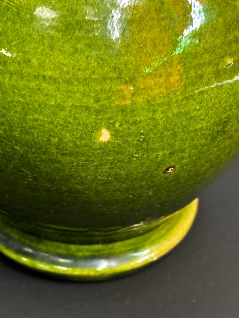 May include: A close-up of a green ceramic bowl with a glossy finish. The bowl has a rounded shape with a yellow rim and a dark base. The green color has variations, with some areas appearing lighter and others darker.