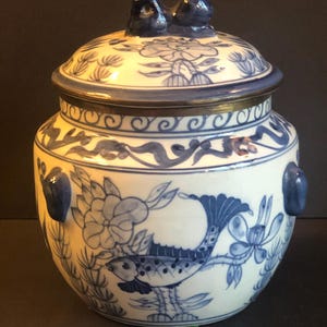 May include: A white and blue ceramic jar with a lid. The jar features a detailed blue floral and fish design. The lid has two blue knobs. The jar has two blue handles on the sides. The jar is a decorative piece.
