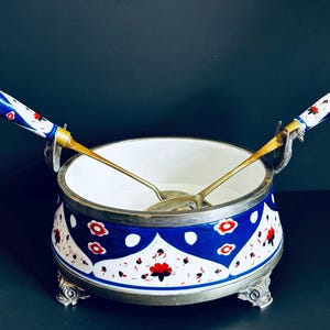 May include: A decorative serving bowl with a white interior and a blue and white exterior design featuring red floral accents. The bowl is paired with two serving spoons with ornate handles. The bowl rests on decorative silver-toned feet.