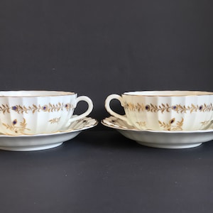 Puede incluir: Dos tazones de sopa de porcelana blanca con ribetes dorados y azules y platillos a juego. Los tazones tienen dos asas y un diseño acanalado.