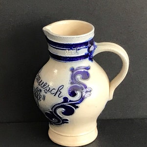 May include: A white ceramic jug with blue and white floral designs and the text "Hausch. Cafe" in blue. The jug has a blue stripe around the top and a handle.