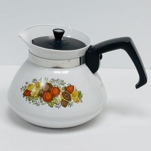 Könnte beinhalten: Weiße Emaille-Teekanne mit schwarzem Griff und Deckel. Die Teekanne hat ein Blumendesign mit orangefarbenen, braunen und gelben Akzenten. Die Teekanne hat ein Edelstahlband in der Mitte.