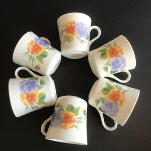 Puede incluir: Seis tazas de cerámica blanca con un diseño floral con pensamientos amarillos, naranjas y morados. Las tazas tienen una forma redondeada y un asa.