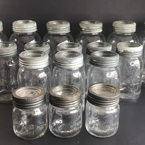 May include: A selection of clear glass preserving jars with silver metal lids. The jars are of various sizes, some taller and narrower, others shorter and wider. Some jars have embossed text. The jars are arranged on a dark surface.