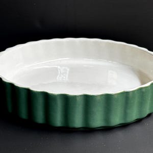 Può includere: Una pirofila rotonda in ceramica con esterno scanalato verde scuro e interno bianco. Il piatto ha un bordo smerlato ed è progettato per cuocere torte, quiche o altri piatti simili. Il piatto è vuoto.