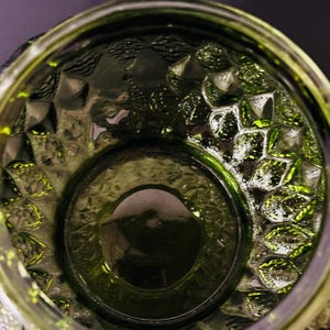 May include: Close-up of a green glass container with a textured, geometric pattern. The container has a wide opening and a thick base. The glass has a glossy finish, reflecting light and creating highlights.