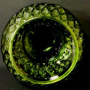 May include: A top-down view of a green glass bowl with a diamond pattern around the rim. The bowl has a circular shape with a deep, dark center. The glass has a glossy finish, reflecting light and creating highlights.