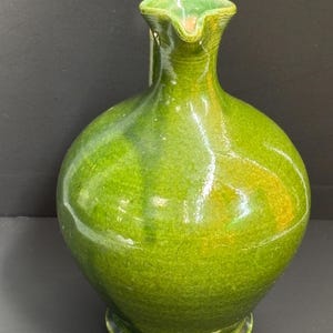 May include: A green ceramic pitcher with a rounded body and a spout. The pitcher has a glossy finish with variations in the green color, and a small handle. The base is a darker shade of green.