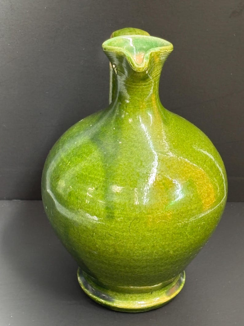 May include: A green ceramic pitcher with a rounded body and a spout. The pitcher has a glossy finish with variations in the green color, and a small handle. The base is a darker shade of green.