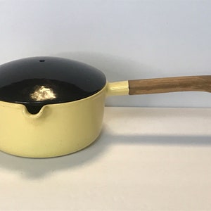 Pode incluir: Uma panela de ferro fundido amarela com uma tampa preta e uma alça de madeira. A panela tem um bico de despejo.