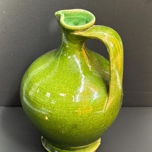 May include: A green ceramic pitcher with a rounded body and a curved handle. The pitcher has a spout and sits on a small, circular base. The glaze has a glossy finish, and the color is a vibrant, bright green.
