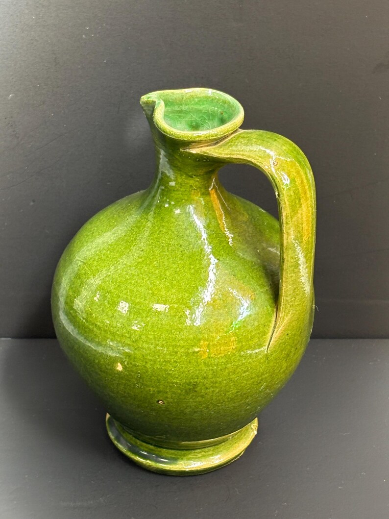 May include: A green ceramic pitcher with a rounded body and a curved handle. The pitcher has a spout and sits on a small, circular base. The glaze has a glossy finish, and the color is a vibrant, bright green.