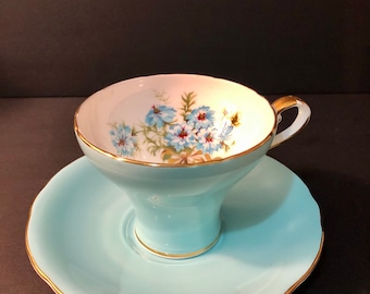 Può includere: Un set da tè composto da una tazza e un piattino di colore azzurro chiaro con un motivo floreale. La tazza e il piattino hanno un bordo dorato.