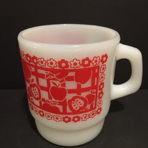 May include: White ceramic mug with a red apple and flower design. The mug has a rounded handle and a white interior.
