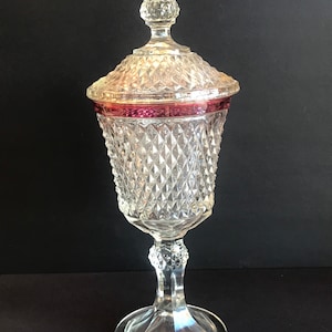 May include: A clear glass candy dish with a diamond pattern and a red band around the top. The dish has a tall, slender base and a domed lid.