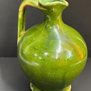 May include: A green ceramic pitcher with a rounded body, a handle, and a spout. The pitcher has a glossy finish and a slightly uneven surface. The handle and the base have a lighter green and yellow color. The pitcher is likely used for serving water or other beverages.